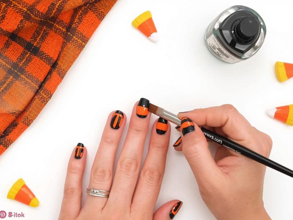Painting glossy black candy corn stripe easy Halloween nail design with orange and yellow stripes.
