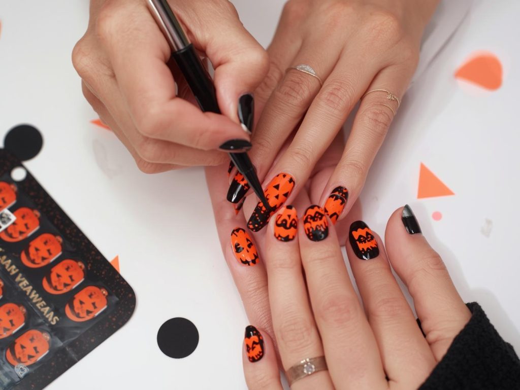 Applying glossy black jack-o’-lantern Kiss Halloween nail design with neon orange patterns.