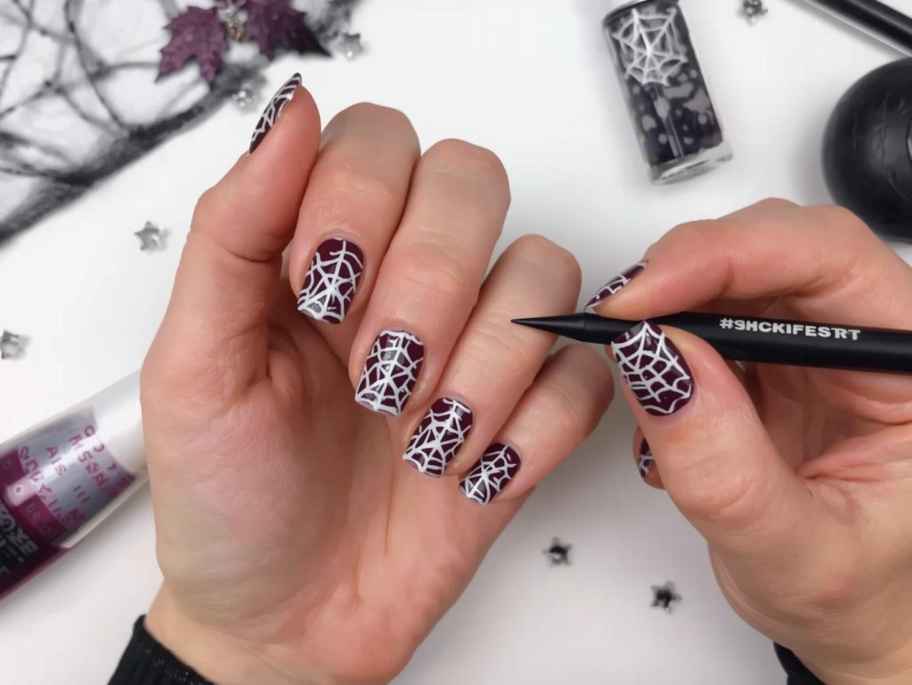 Applying deep plum cobweb short Halloween nail design with white lines.
